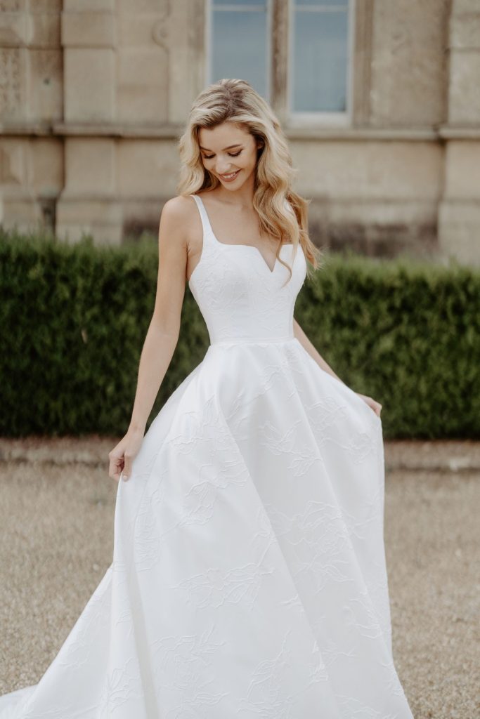 This cloque statement gown, the Suzanne Neville Bloomsbury, in incredibly structured. It has a corseted bodice featuring a modern square neckline that is contoured into a striking plunge, straps and an A-line full skirt with pockets and buttons of course. Find your dream wedding dress at Your Dream Bridal near Boston.
