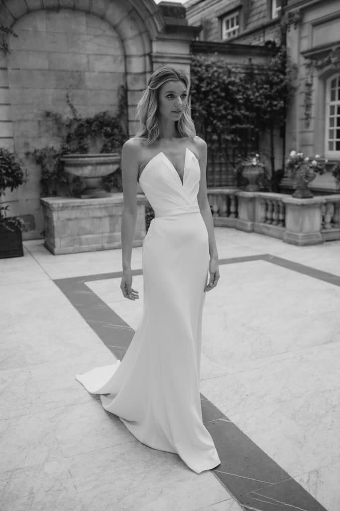 Strapless Suzanne Neville Caccini wedding dress has a gorgeous plunge neckline with corseted bodice, satin detailing and satin waist belt with a softly fitted crepe skirt. Find your wedding dress at Your Dream Bridal in near Boston.