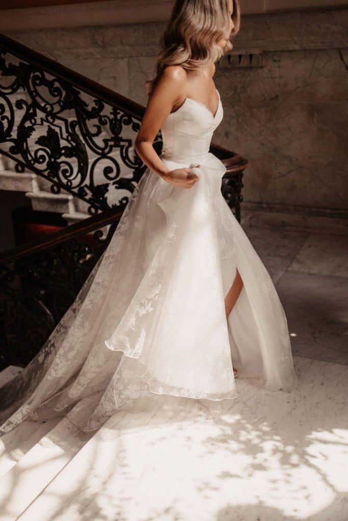 The breathtaking Suzanne Neville Rossini wedding dress has a gorgeous wrapped strapless neckline in embroidered organza with a corseted bodice and soft flowing full A-line skirt with hidden slit.